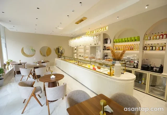 The Castle Patisserie-nin işıqlı zalı, şirniyyat vitrini, tağlı rəfləri və ağ çiçəkli dekoru