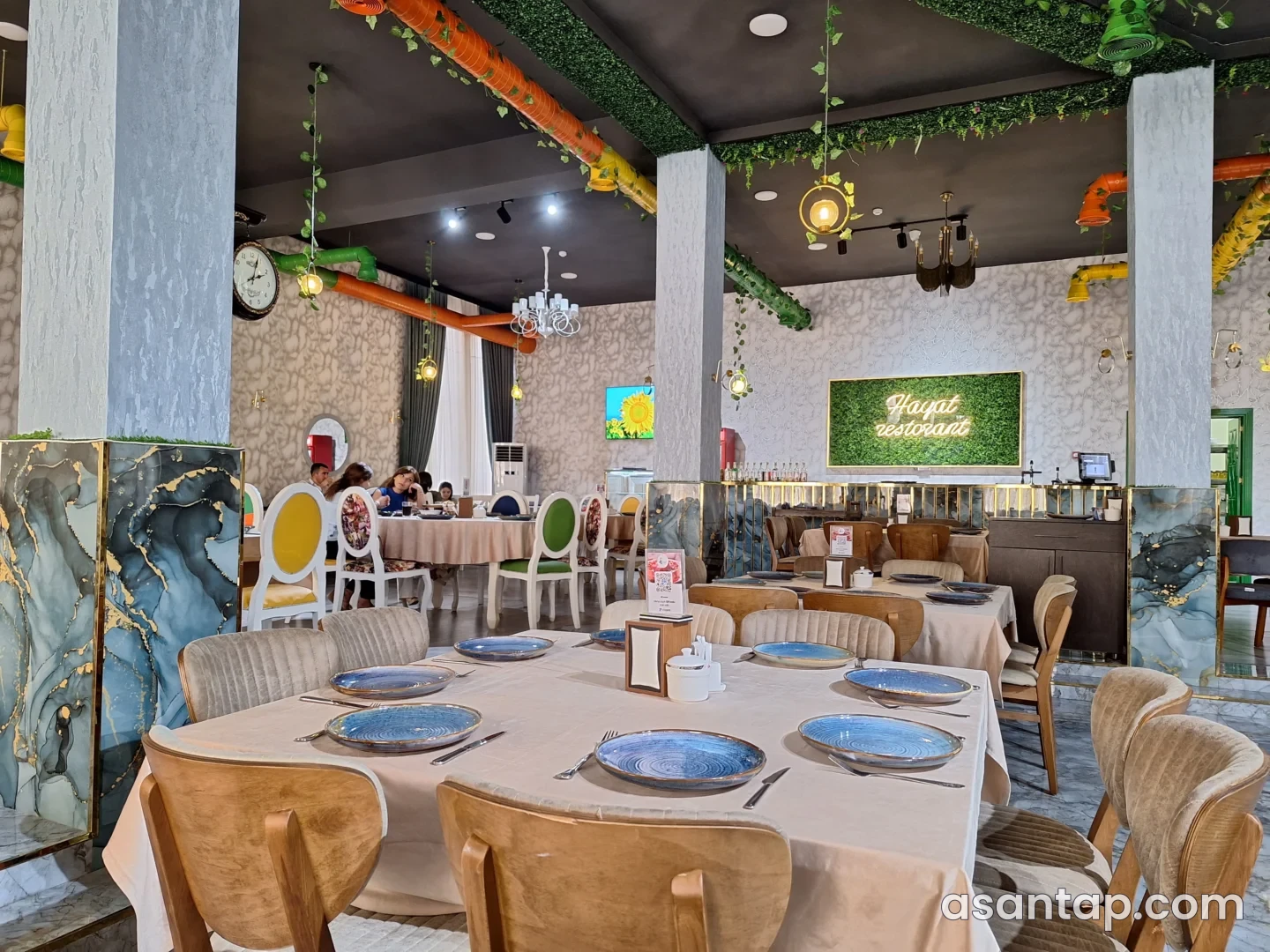 Hayat Restoranının adı yazılmış yaşıl divar lövhəsi, rəngli tavan boruları və süfrə açılmış masalar olan zal