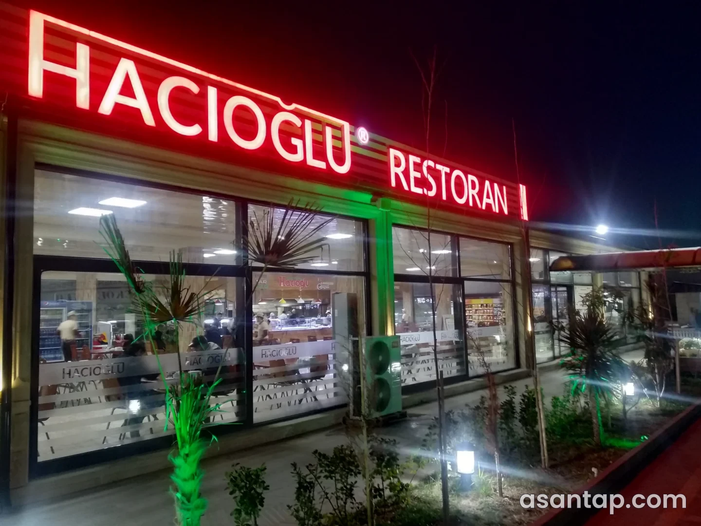 Salyanda Hacıoğlu Restoran II-nin işıqlı yazısı, şüşəli fasadı və qarşısındakı yaşıllıq