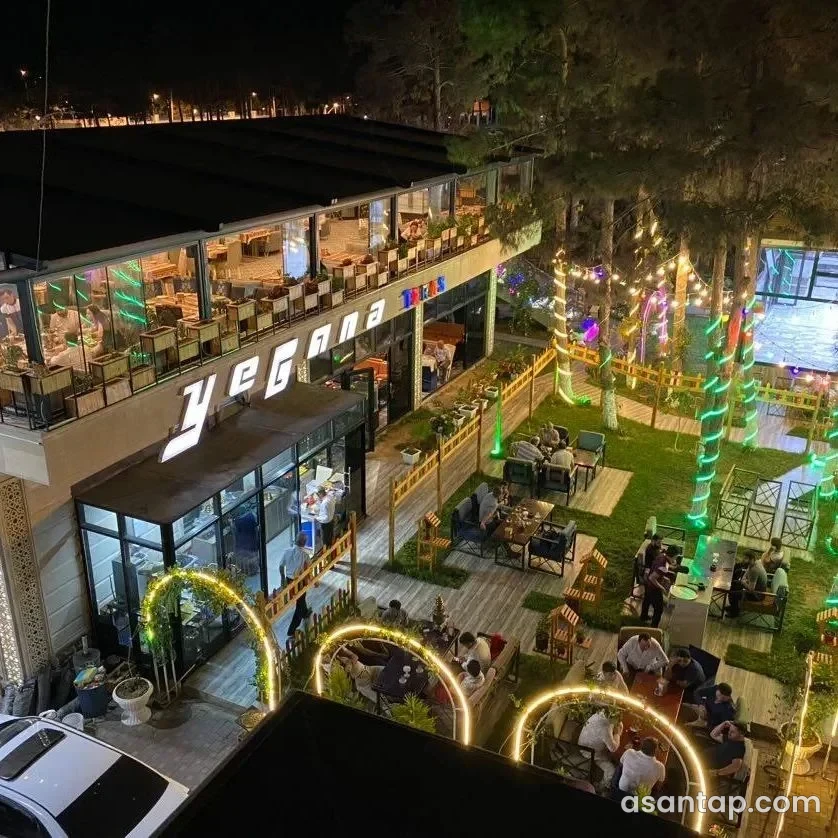 Yeganə Teras restoranının işıqlı həyəti, ağaclar arasındakı masaları və Yeganə yazılı yuxarı terrası