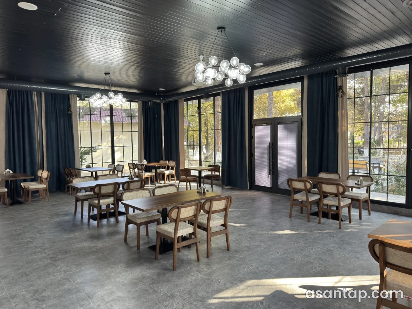 Qar Dənəsi restoranının iri pəncərəli zalı, taxta masaları və park mənzərəsi