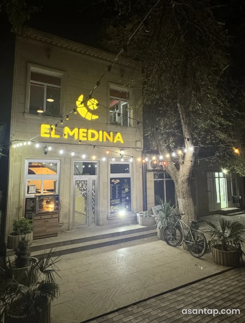 Şabranda sarı El Medina lövhəsi, şüşəli giriş və ağac qarşısındakı işıqlı restoran fasadı