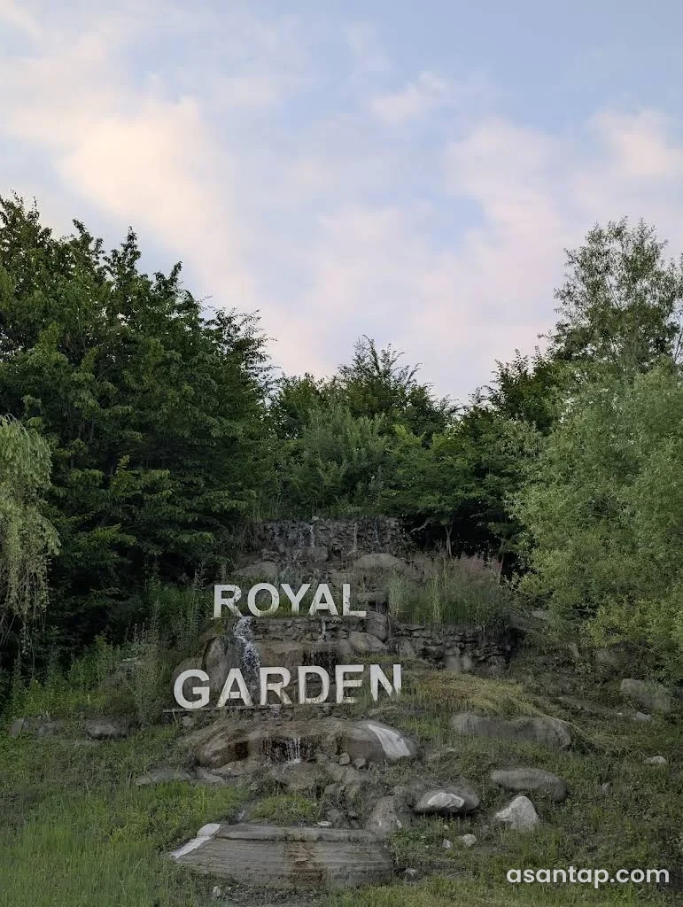 Royal Garden restoranının ağ hərflərlə adı yazılmış daşlı su kaskadı və bağ yaşıllığı