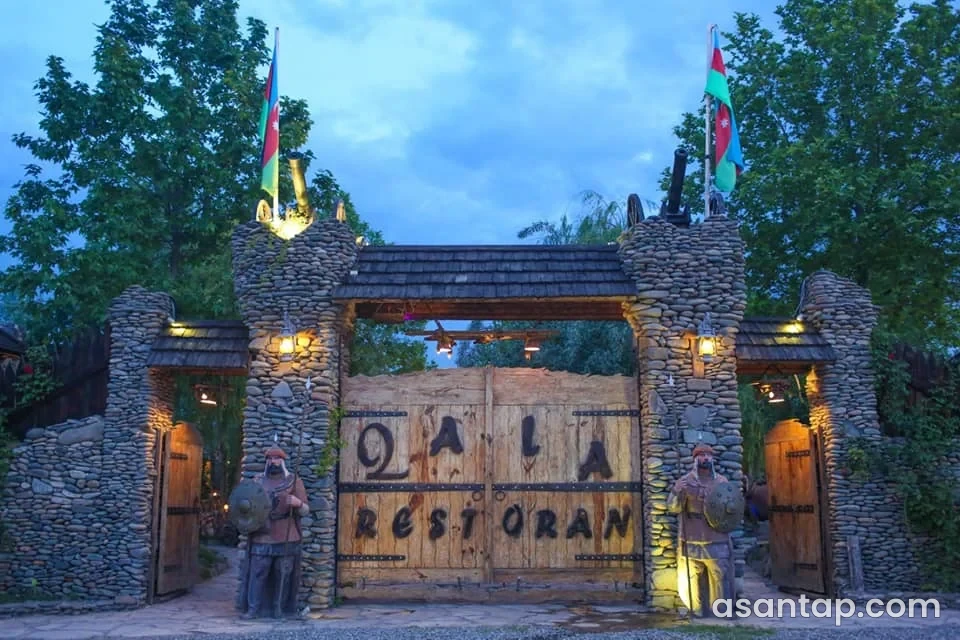 Qala restoranının daş qülləli, taxta qapılı və mühafizəçi fiqurlu imzalı girişi