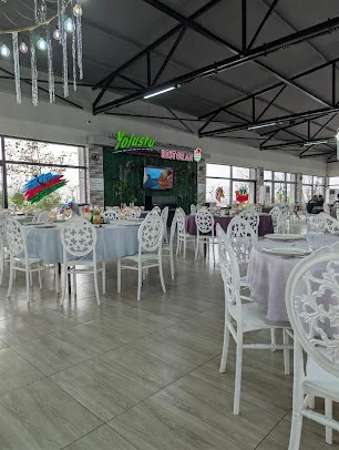 Yolüstü Restoran yazısı olan yaşıl divar, qara metal tavan fermaları, ağ stullar və örtüklü masalarla qurulmuş salon