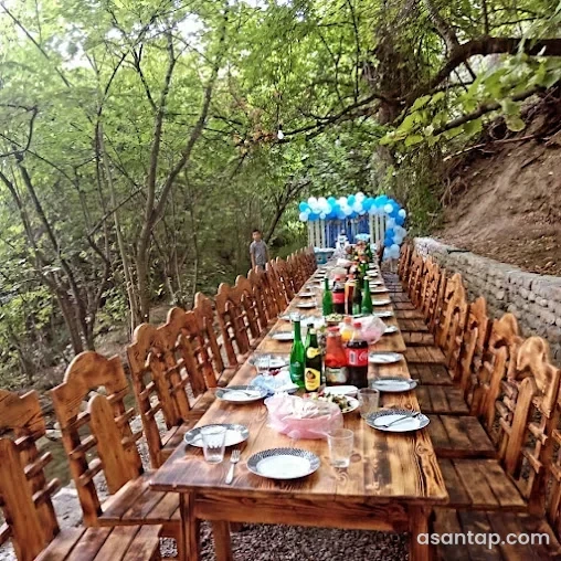 Şabran Soyuq Bulaq restoranında ağacların altında uzun taxta süfrə, oyma kürəklikli stullar və daş istinad divarı