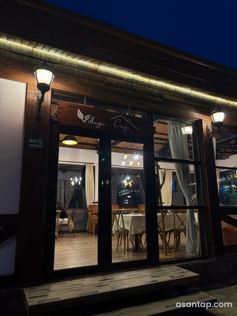 Village Cafe yazılı şüşəli giriş, taxta stullar və ağ örtüklü masalar