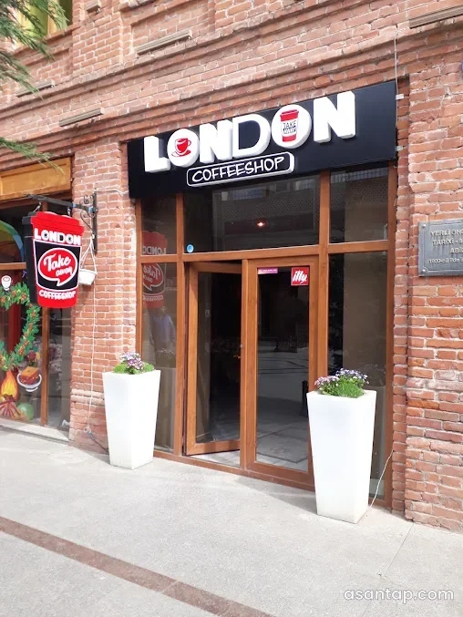 London CoffeeShop-un qırmızı kərpic fasadı, qara-ağ yazılı lövhəsi, şüşəli qəhvəyi qapısı və ağ gül dibçəkləri