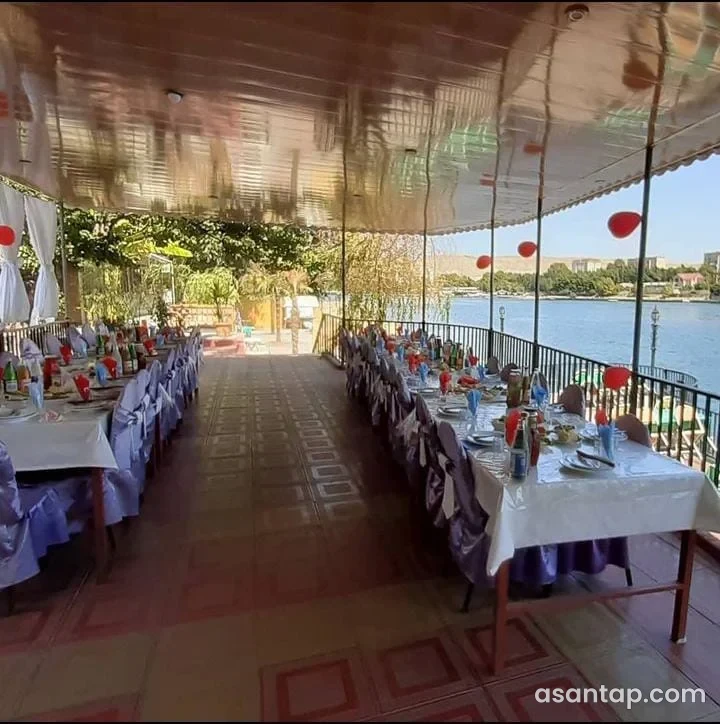 Sərin restoranının Kürə baxan üstüörtülü sahəsində ağ süfrəli uzun masalar və çay mənzərəsi