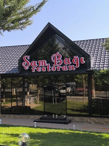 Şam Bağı restoranının şüşəli üçbucaq giriş fasadı, ad lövhəsi və ağaclı yaşıllıq sahəsi
