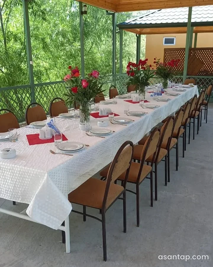 Görüş Yeri ailəvi restoranının örtülü bağ sahəsində ağ örtüklü uzun süfrə, çiçəklər və yaşıl qəfəsli məhəccər