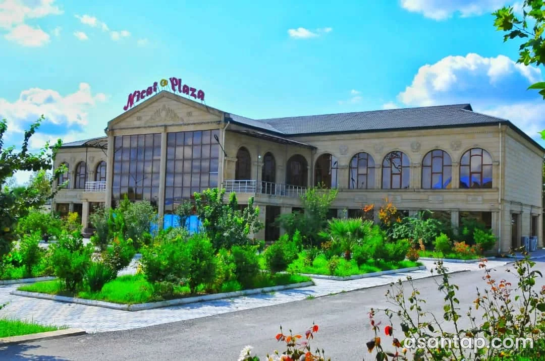 Nicat Plaza yazılı şüşəli mərkəzi fasad, tağlı pəncərələr və qarşıdakı yaşıllıq